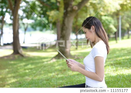 Young woman in a park wearing casual sitting on a mat using mobile device tablet iPad 11155177