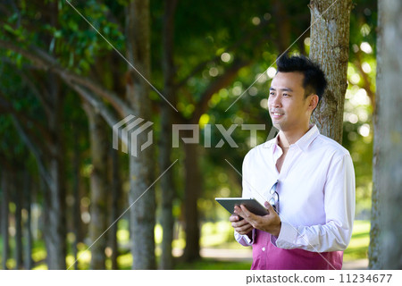Young man in causal leaning in front of a tree using mobile device tablet iPad Young man in causal leaning in front of a tree using mobile device tablet iPad 11234677