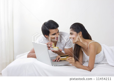 Young man opening ring box with woman shopping online with credit card in bed Young man opening ring box with woman shopping online with credit card in bed 11454211