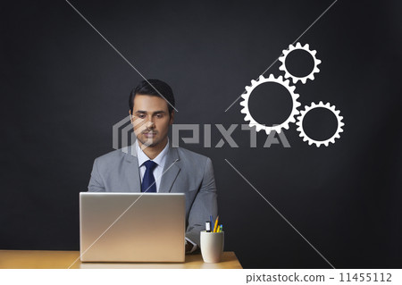 Digitally generated image of businessman using laptop at office desk with gears 11455112