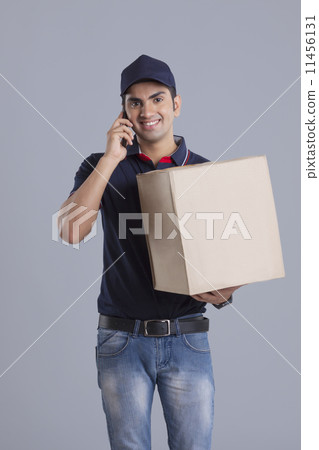 Portrait of delivery man carrying package while answering mobile phone against gray background 11456131
