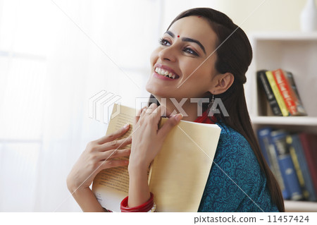 Smiling young woman holding book in living room Smiling young woman holding book in living room 11457424