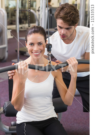 Fit brunette using weights machine for arms with trainer helping 11533513