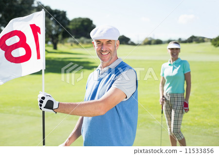 Happy golfer holding eighteenth hole flag with partner behind hi Happy golfer holding eighteenth hole flag with partner behind hi 11533856
