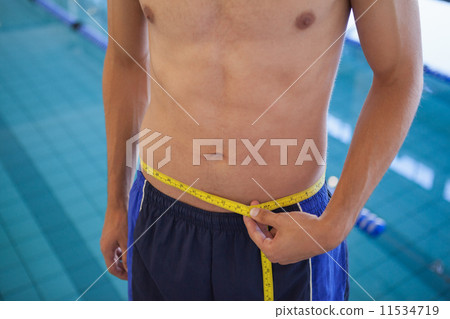 Fit man in swimming trunks standing by the pool measuring his wa 11534719