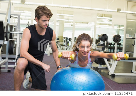 Trainer watching client balance on exercise ball with dumbbells Trainer watching client balance on exercise ball with dumbbells 11535471