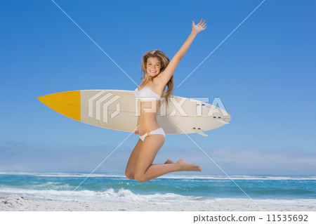 Smiling surfer girl holding her surfboard and jumping on the bea 11535692