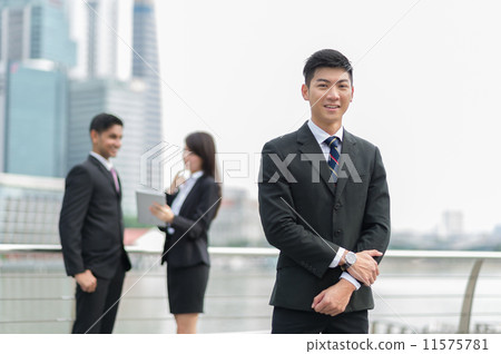 A young and dynamic Chinese businessman poses in the foreground while his co-workers standing behind A young and dynamic Chinese businessman poses in the foreground while his co-workers standing behind 11575781