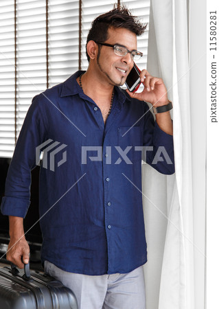 Cheerful Asian man chatting on the phone, hand holding luggage bag, indoors 11580281
