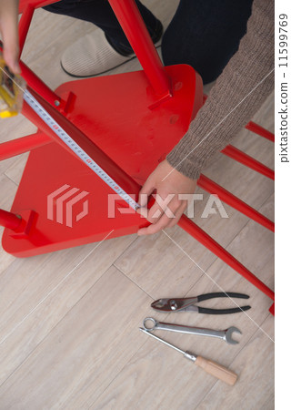 A woman uses a measuring tape to measure the assembly of a red chair. 11599769
