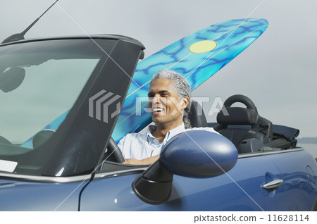 Senior man driving a convertible with a surfboard in it  11628114