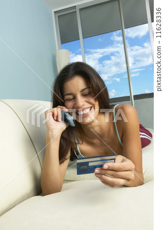 Hispanic woman laying on sofa using cell phone with credit card 11628336