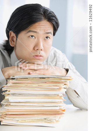 Asian businessman leaning on stack of paperwork Asian businessman leaning on stack of paperwork 11629492