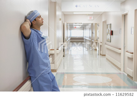 Indian male doctor in scrubs leaning against wall in hospital Indian male doctor in scrubs leaning against wall in hospital 11629582
