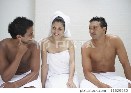 Two men and a woman in towels sitting on bench in bathroom 11629980