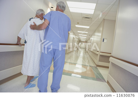 Rear view of male nurse helping senior patient walk in corridor 11630876