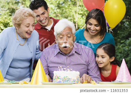Senior Hispanic man blowing out birthday candles with family in park 11631353