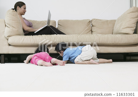 Mother working on laptop while children look under couch Mother working on laptop while children look under couch 11633301