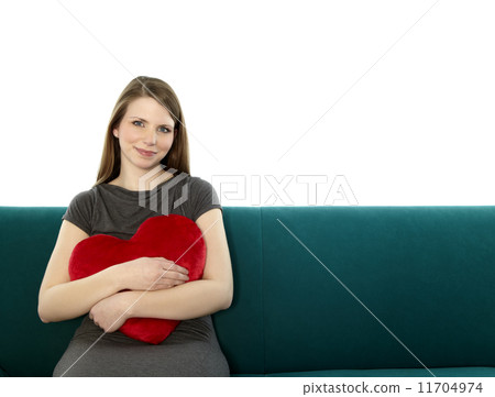Beautyful young and smiling woman sitting on a sofa embracing heart shaped pillow with her hands Beautyful young and smiling woman sitting on a sofa embracing heart shaped pillow with her hands 11704974