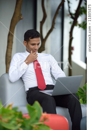 Successful young Indian corporate executive using laptop sitting down along promenade Successful young Indian corporate executive using laptop sitting down along promenade 11805875