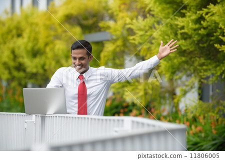 Successful young Indian corporate executive rejoicing while using laptop along promenade 11806005