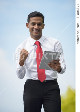 Successful young Indian corporate executive victory using iPad mobile device tablet along promenade Successful young Indian corporate executive victory using iPad mobile device tablet along promenade 11806177