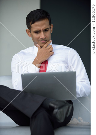 Successful young Indian corporate executive using laptop sitting down along promenade Successful young Indian corporate executive using laptop sitting down along promenade 11806179