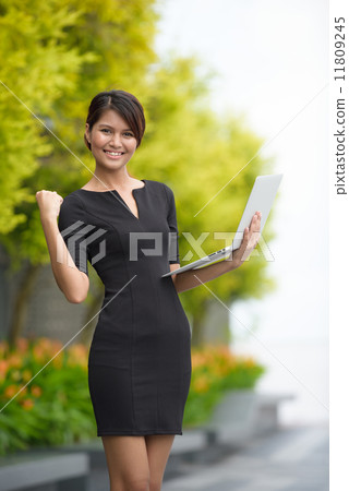 Victorious successful young Malay business woman using laptop along promenade Victorious successful young Malay business woman using laptop along promenade 11809245