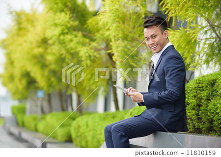 Successful young Chinese businessman using iPad mobile device tablet sitting along promenade Successful young Chinese businessman using iPad mobile device tablet sitting along promenade 11810159