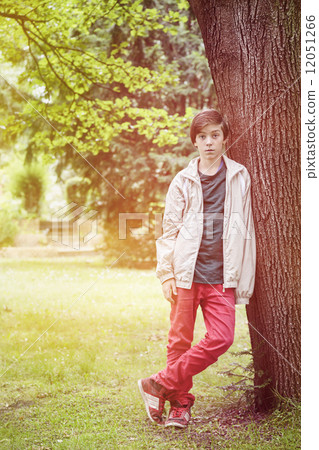 teenager boy leaning against a tree in a park 12051266
