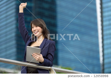 Victorious young Chinese business woman with iPad punching her fist in the air in front of building Victorious young Chinese business woman with iPad punching her fist in the air in front of building 12058998