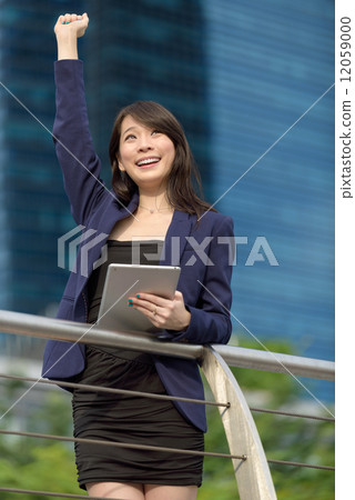 Victorious young Chinese business woman with iPad punching her fist in the air in front of building 12059000