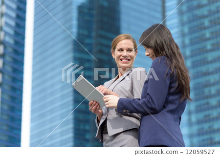 Two young successful business women with tablet iPad in discussion in front of glass office building 12059527