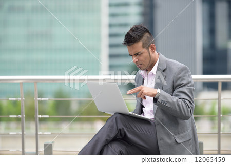One successful young Malay business man using laptop along promenade in front of office building One successful young Malay business man using laptop along promenade in front of office building 12065459