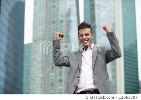 Victorious young successful Malay business man punching fist in the air in front of office buildings Victorious young successful Malay business man punching fist in the air in front of office buildings 12065597