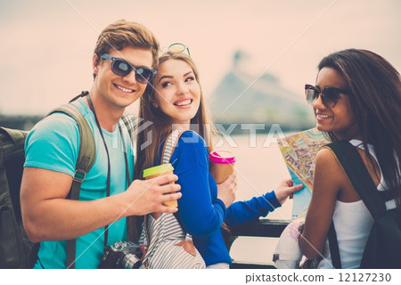 Multi-ethnic friends tourists with map and coffee cups near river in a city Multi-ethnic friends tourists with map and coffee cups near river in a city 12127230