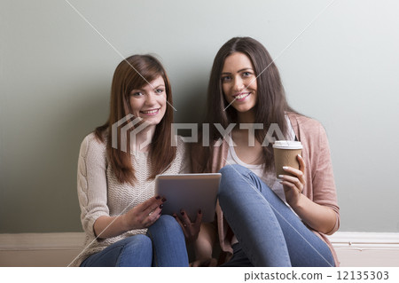 Two young female friends sitting at home, reading a tablet pc Two young female friends sitting at home, reading a tablet pc 12135303