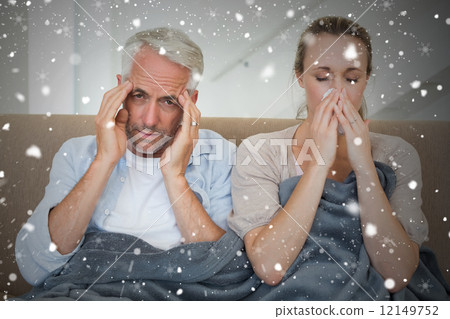 Composite image of sick couple sitting on the couch under a blan 12149752