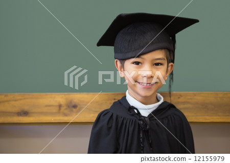 Cute pupil in graduation robe smiling at camera in classroom 12155979