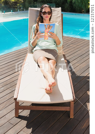 Woman reading book on sun lounger by pool 12157051