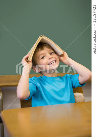 Happy pupil holding book on his head at desk 12157422