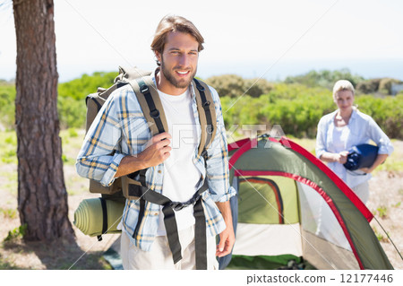 Attractive man smiling at camera while partner pitches tent Attractive man smiling at camera while partner pitches tent 12177446