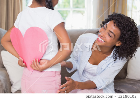 Pretty mother sitting on couch with daughter hiding heart card Pretty mother sitting on couch with daughter hiding heart card 12180102