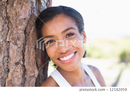 Fit woman leaning against tree smiling at camera Fit woman leaning against tree smiling at camera 12182059