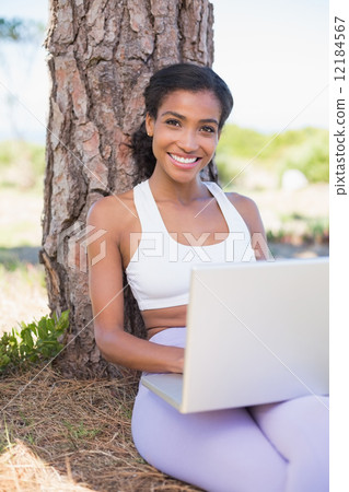 Fit woman sitting against tree using laptop 12184567