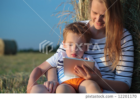 Mother and son with pad sitting by hay roll in the field 12201170