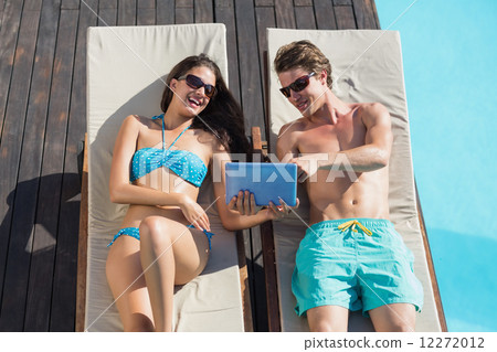 Couple using digital tablet on sun loungers by swimming pool 12272012