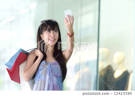 Young chinese woman shopping doing selfie in front of glass display window  12423594