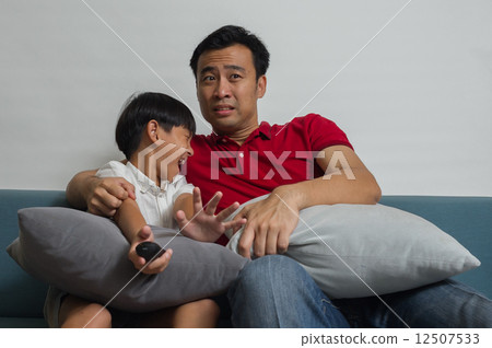 Asian Father and son are terrified / afraid watching a scary movie on a sofa in the living room 12507533