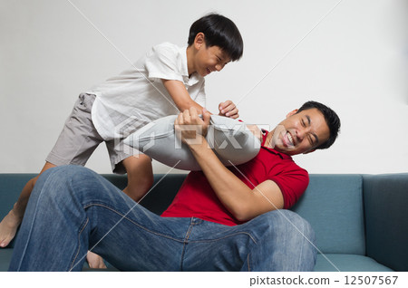 Asian boy is playing - pillow fight - with his dad / father on the sofa in the living room 12507567